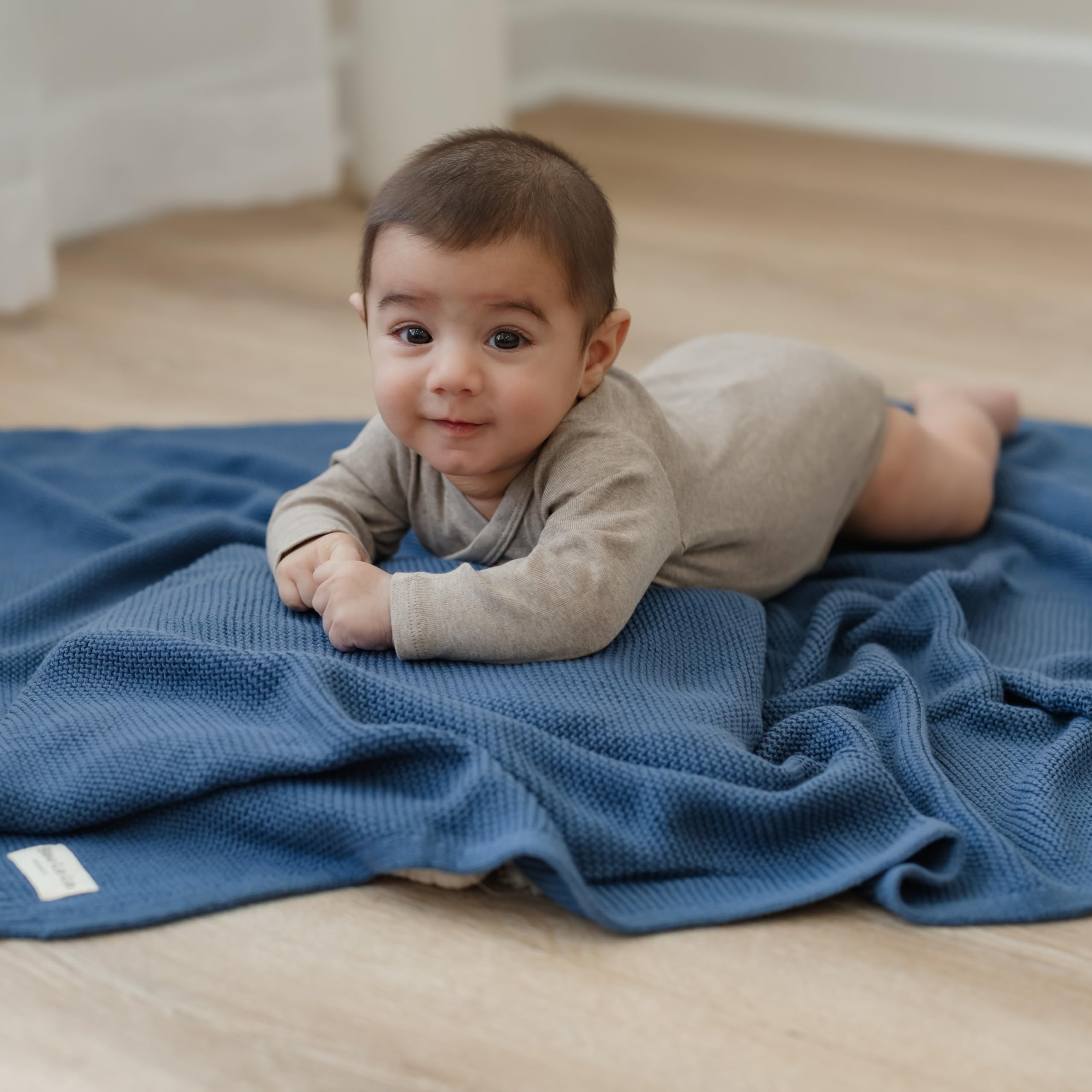 Baby head-on view of denim blue Heritage Knit swaddle on soft surface