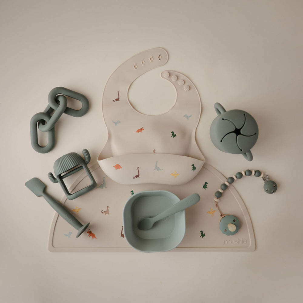 Mushie dinosaur bib pairs with placemats and tableware for meals.