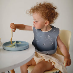 Mushie silicone baby bib deep pocket closeup, boats design, helps catch crumbs.